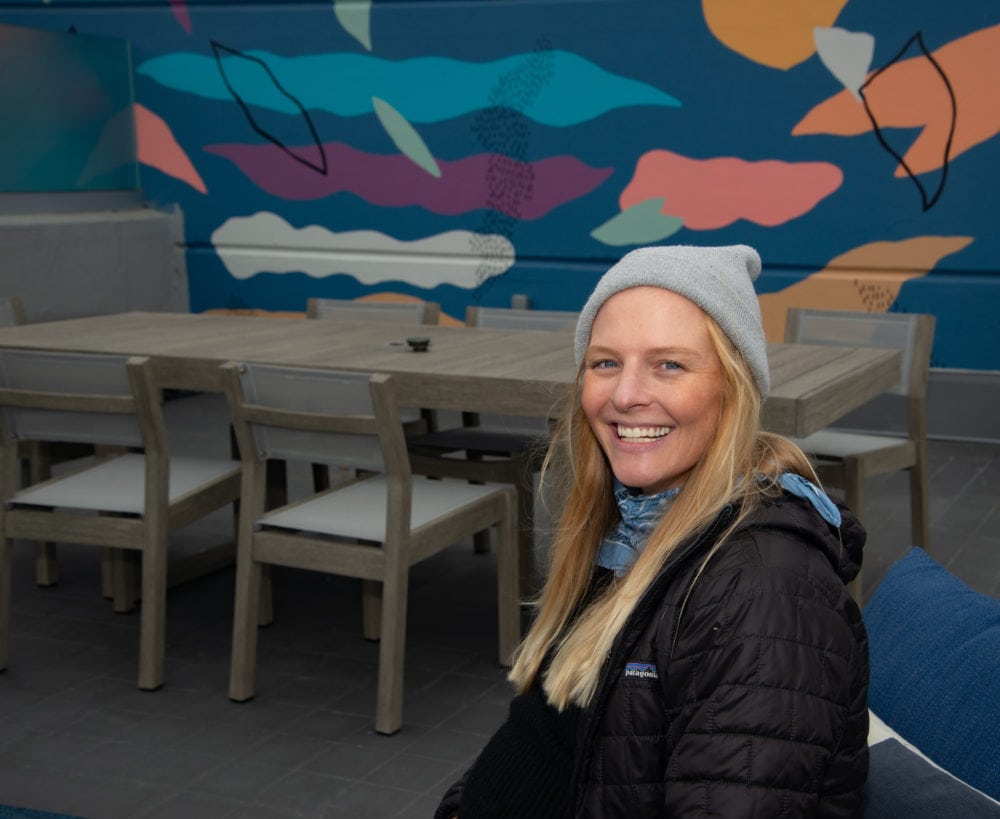 Picture of Priscillla Witte wearing jacket and beanie, sitting in front of Sunset Deck mural