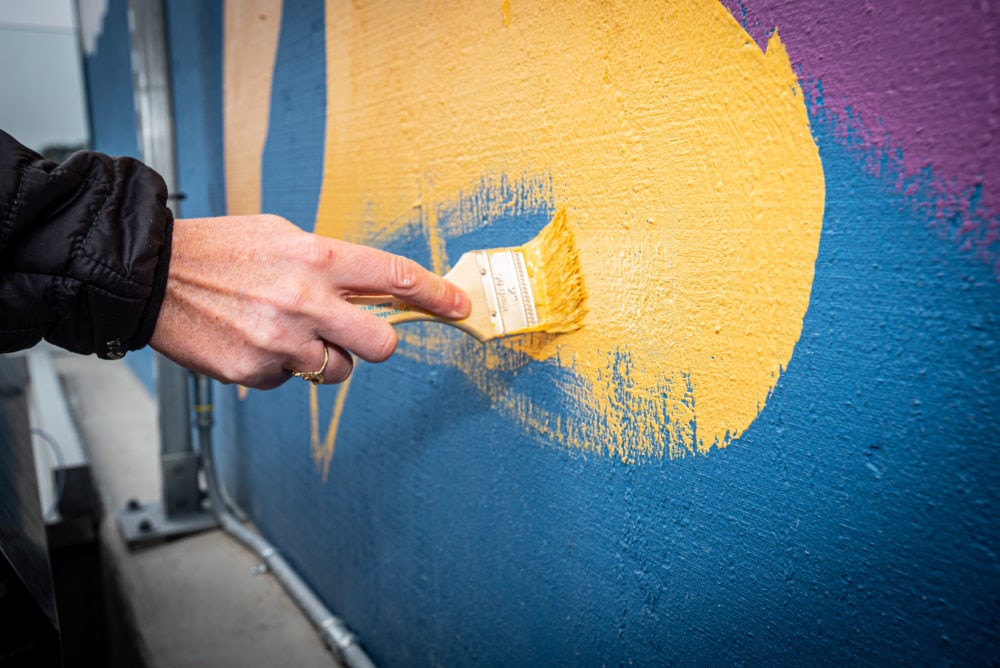 Close up shot of mural being painted, hand holding paintbrush dipped in bright yellow