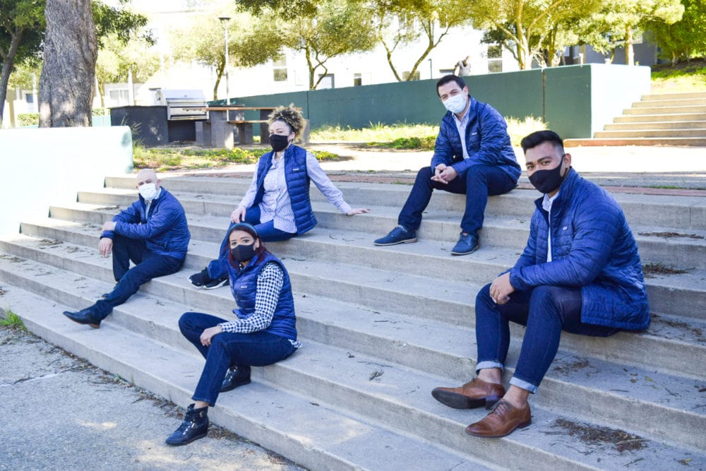 5 Maximus employees sitting on steps with. masks and blue jackets